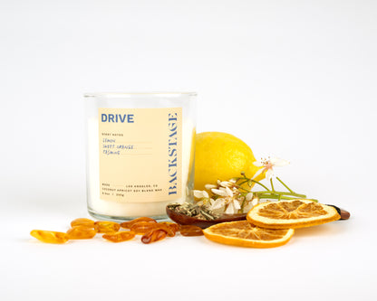 A lit white candle with a label that reads 'DRIVE' and 'Backstage', surrounded by a lemon, orange slices, amber, and jasmine flower