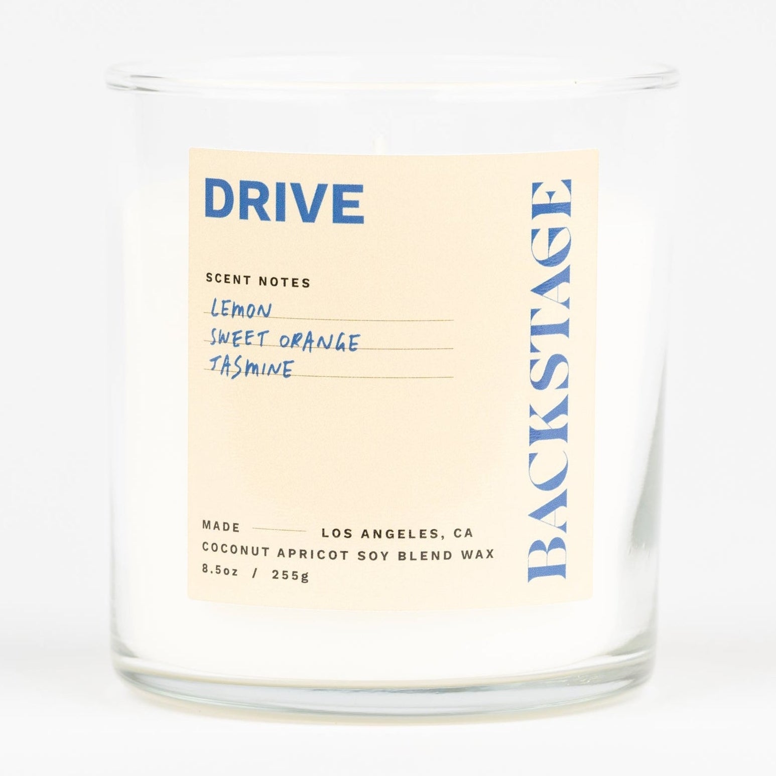 A white candle with a label that reads 'DRIVE' and 'Backstage', with notes of Lemon, Tea, Jasmine, Lemongrass, Amber, and Musk.