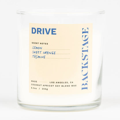 A white candle with a label that reads 'DRIVE' and 'Backstage', with notes of Lemon, Tea, Jasmine, Lemongrass, Amber, and Musk.