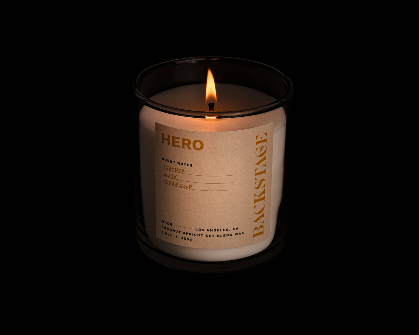 A lit white candle with a label that reads 'HERO' and 'Backstage', with notes of Leather, Musk, and Cedarwood