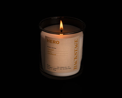 A lit white candle with a label that reads 'HERO' and 'Backstage', with notes of Leather, Musk, and Cedarwood