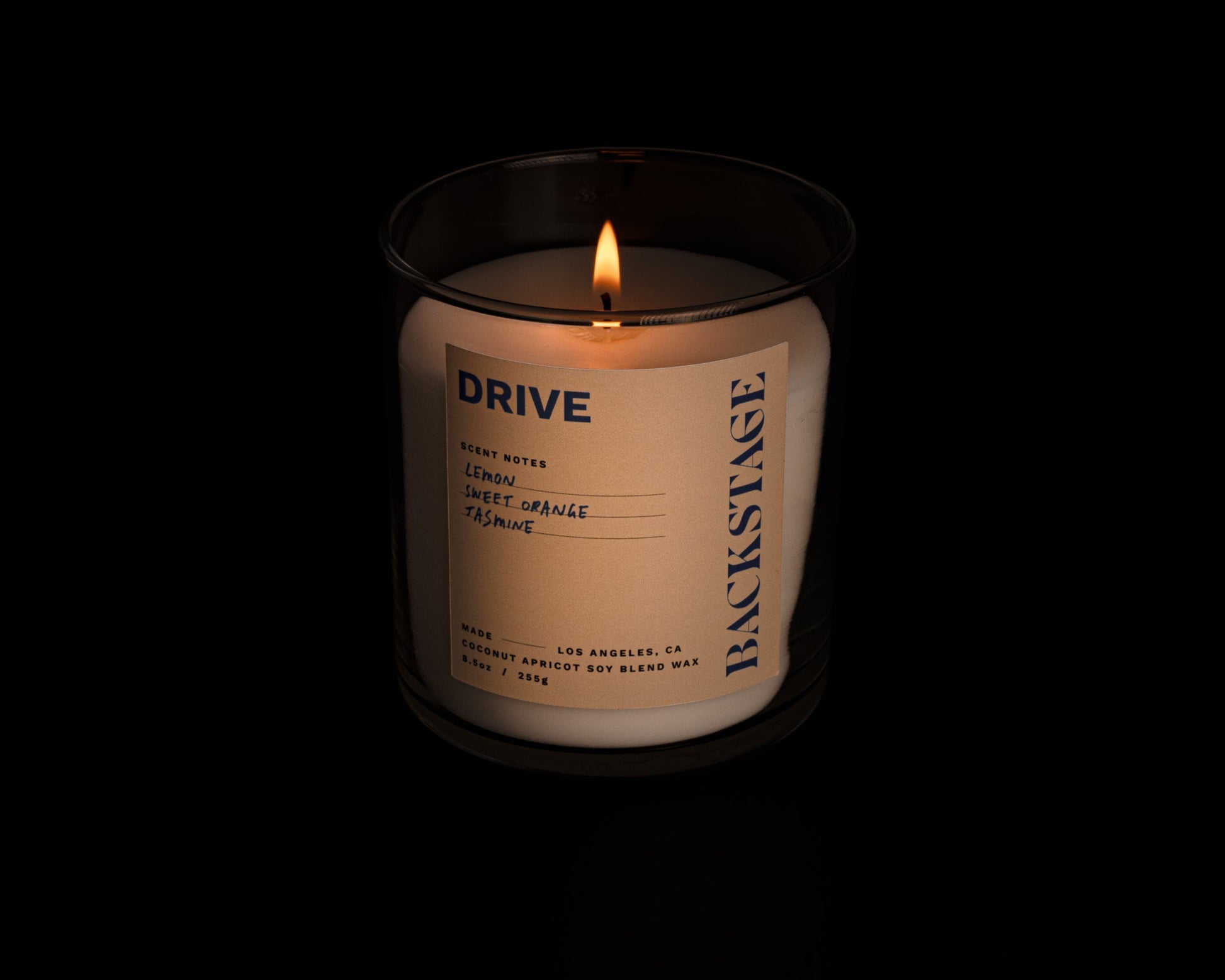 A lit white candle with a label that reads 'DRIVE' and 'Backstage', with notes of Lemon, Tea, Jasmine, Lemongrass, Amber, and Musk.